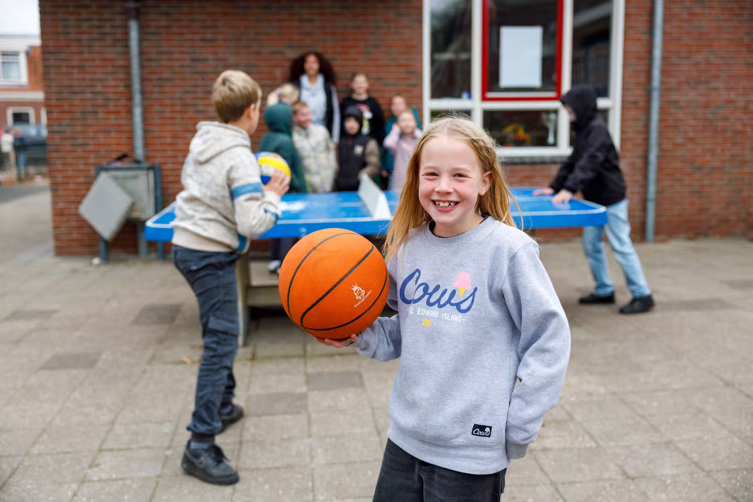 Lachend meisje met een basketbal, kinderen spelen pingpong en staan buiten bij een gebouw.
