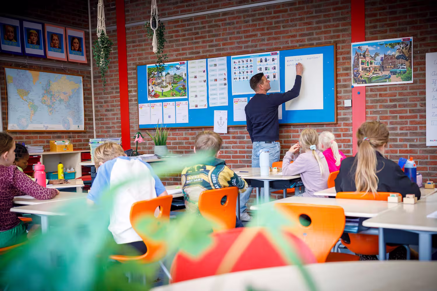 Leraar schrijft op bord terwijl kinderen met oranje stoelen in een klaslokaal zitten.