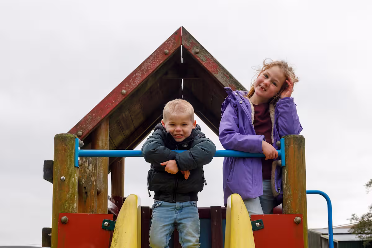 Twee kinderen leunen over een blauwe balk op een houten speeltoestel buiten.