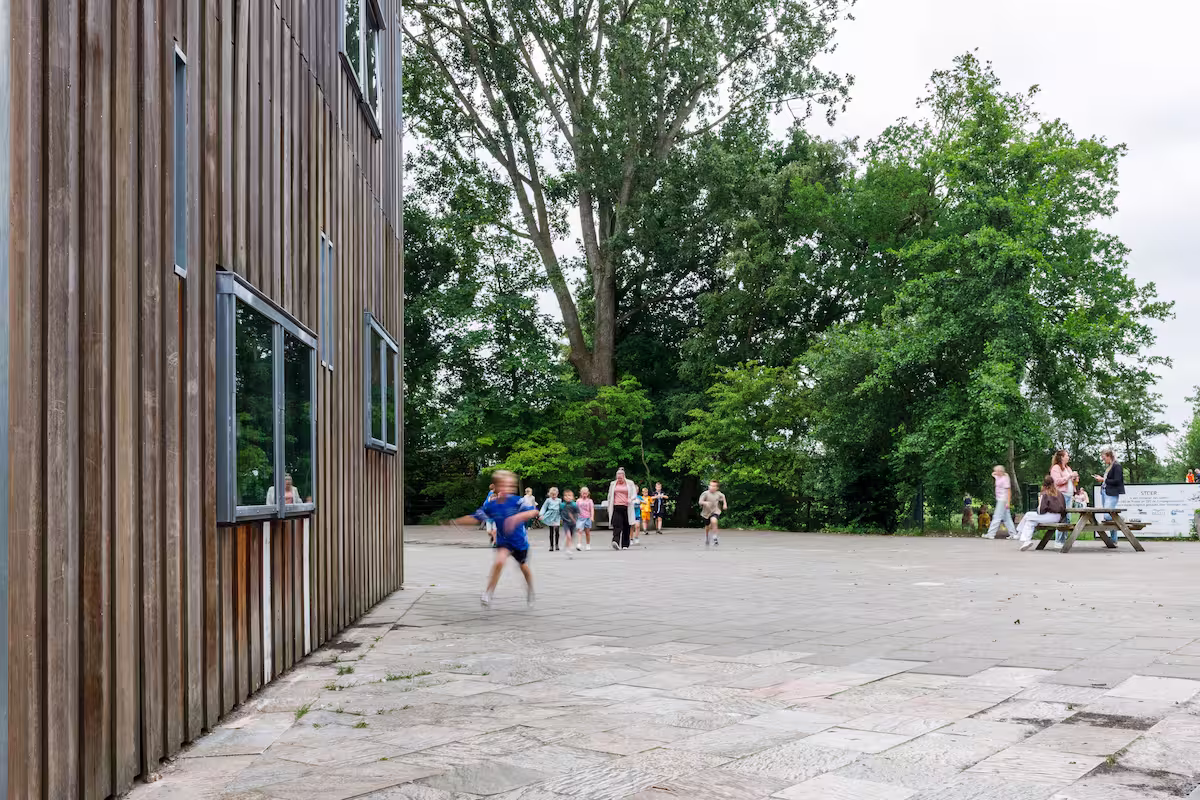 Kinderen rennen en spelen buiten op een plein naast een houten gebouw met bomen op de achtergrond.