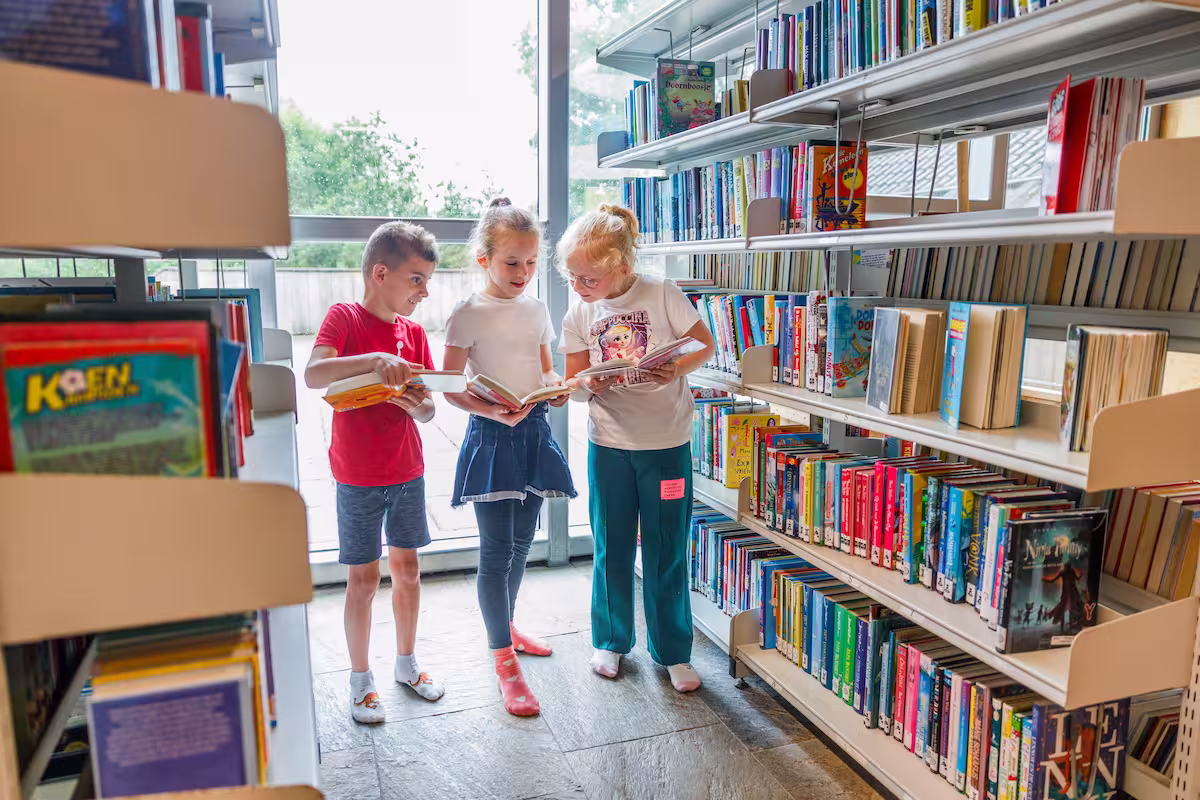 Drie kinderen staan in bibliotheekgang, lezen samen boeken uit de kinderboekenplank.