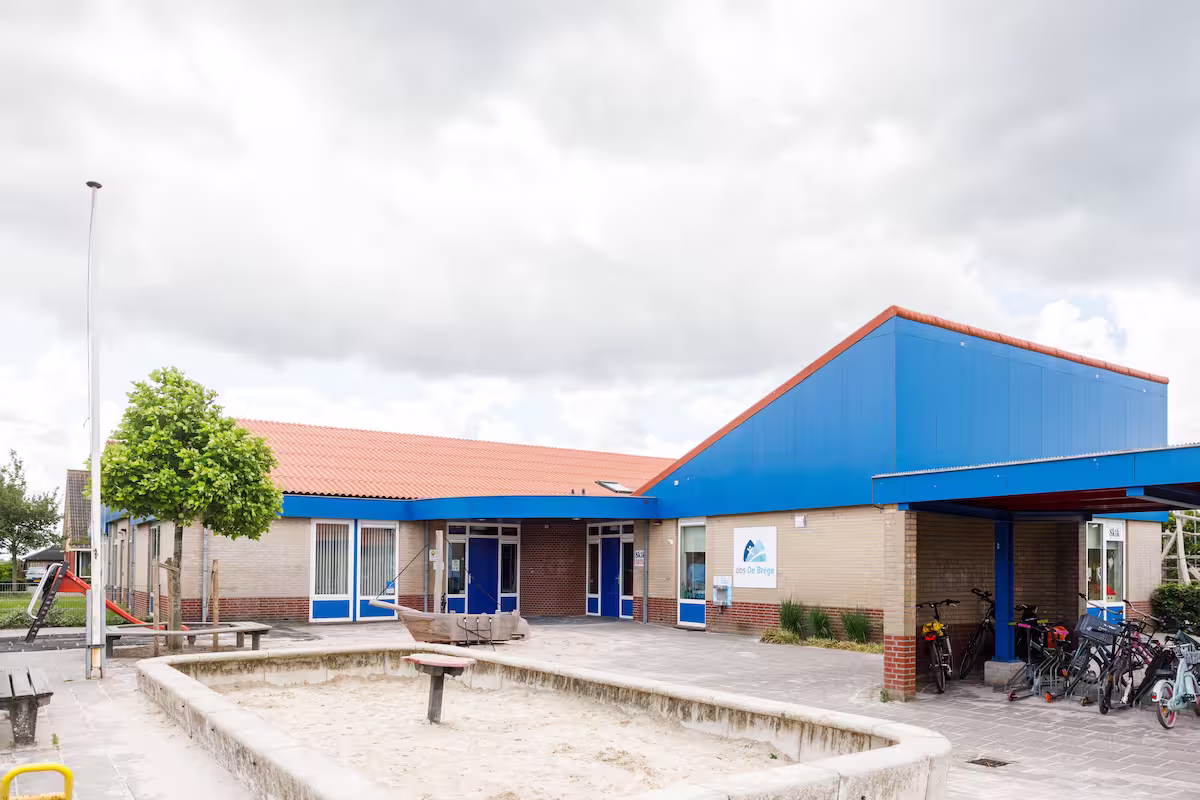 Schoolgebouw met zandbak, fietsenstalling en rood dak onder bewolkte hemel.