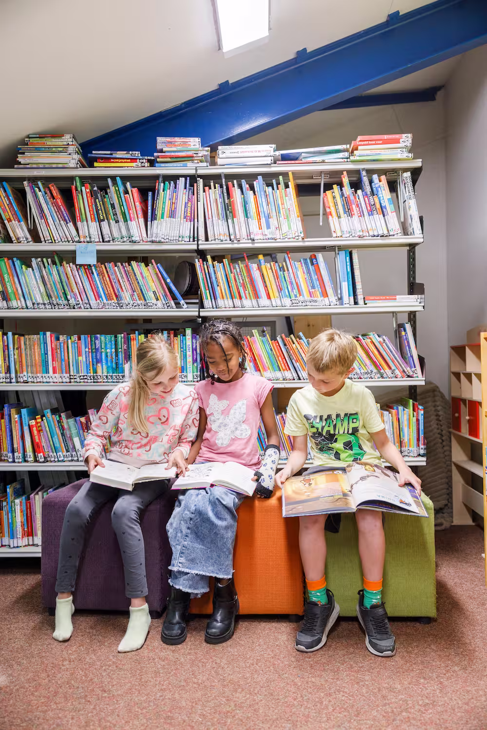 Drie kinderen zitten in een bibliotheek leest boeken op gekleurde banken voor boekenplanken.
