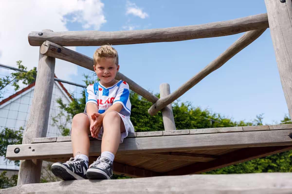 Kind zit lachend op houten speeltoestel buiten onder blauwe lucht.