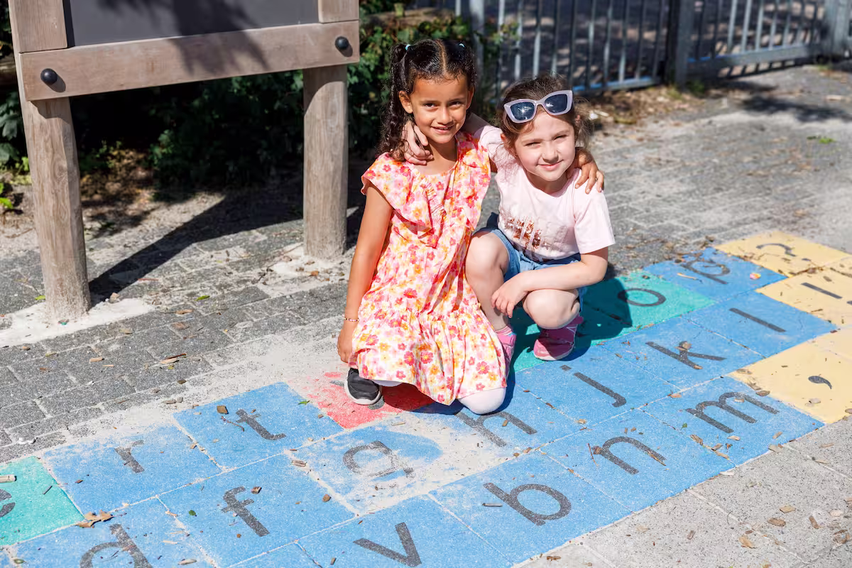 Twee jonge meisjes knielen en lachen samen op een kleurrijk speelplein alfabetspel buiten.