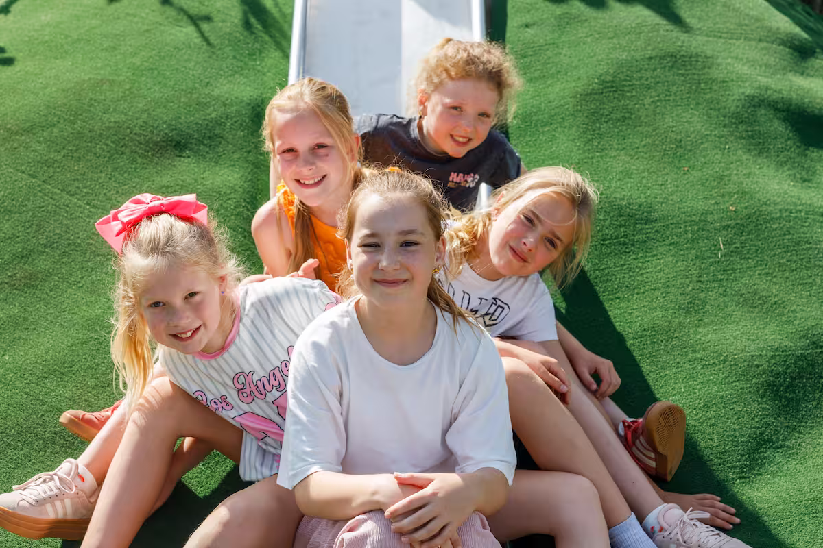 Vijf lachende meisjes zitten op kunstgras voor een glijbaan buiten in de zon.