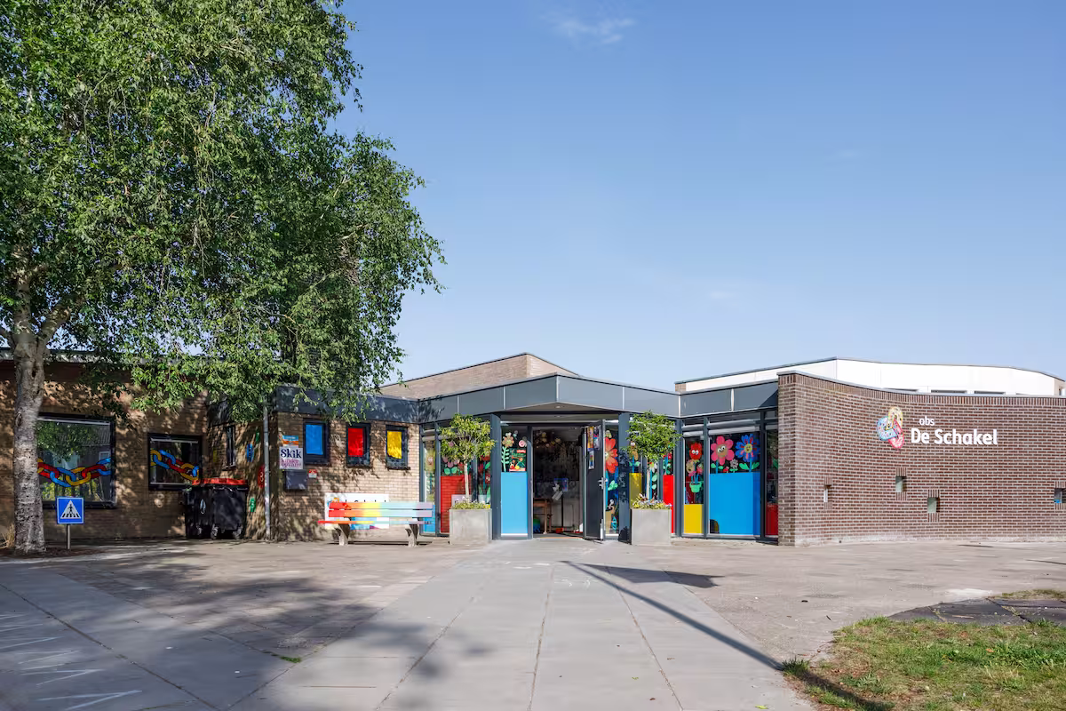 Vooraanzicht van basisschool De Schakel met kleurrijke ramen en een regenboogbank onder een boom.