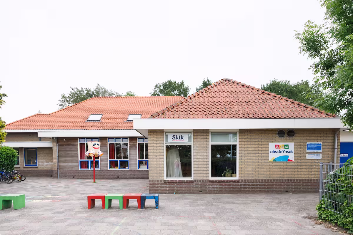 Schoolgebouw met rode dakpannen, kleurrijke stoelen buiten en een speelplaats met fietsen.