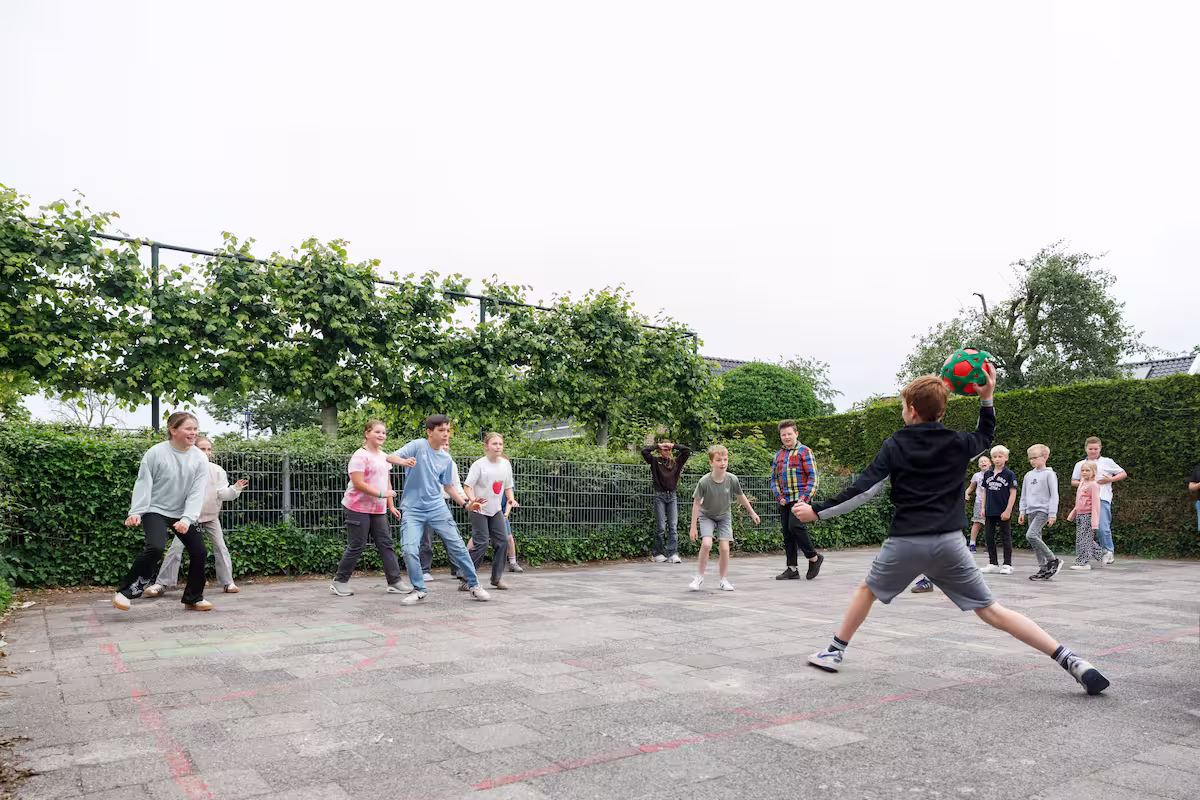 Kinderen spelen een balspel buiten op een betegeld veld met groene heggen en bomen op de achtergrond.