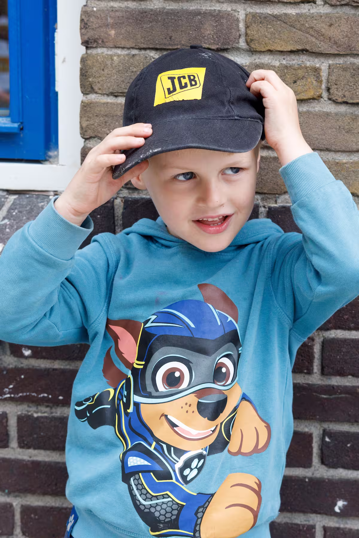 Jongen met zwarte JCB-pet en blauw shirt met getekende hond tegen bakstenen muur.
