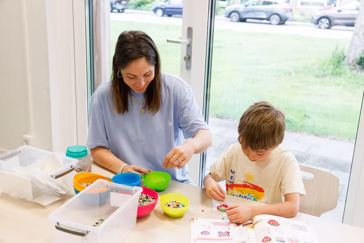 Vrouw en jongen maken kralenfiguren aan tafel met kleurrijke bakken kralen en instructieboek.