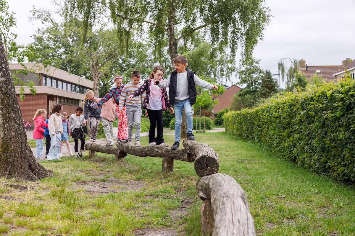 Kinderen balanceren op houten balk buiten in een groene buurt tijdens een schoolactiviteit.