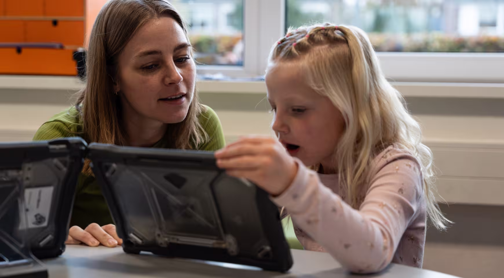 Vrouw begeleidt jong meisje dat op een tablet in een klaslokaal kijkt.