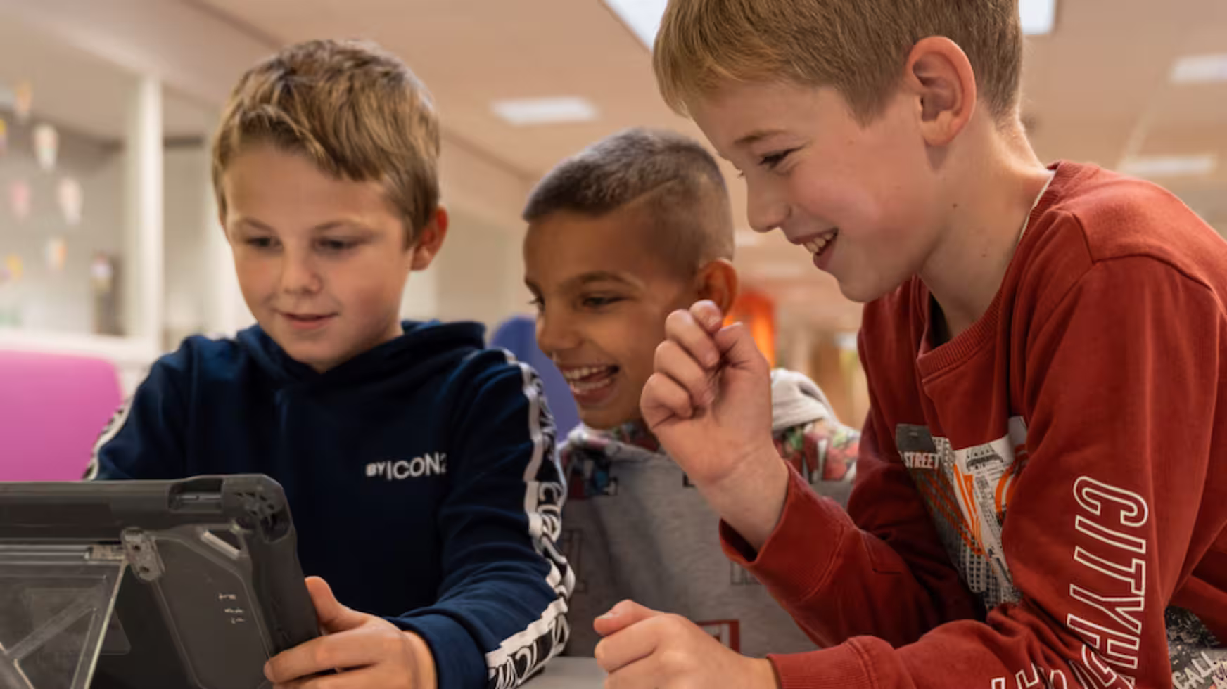 Drie jongens lachen en kijken samen naar een tablet in een binnenruimte.