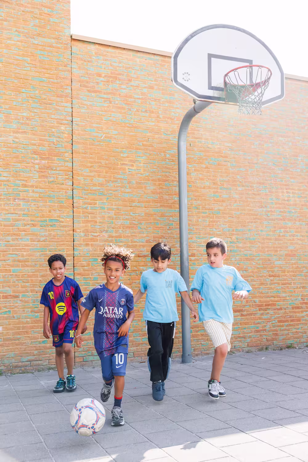 Vier jongens spelen samen buiten met een voetbal bij een basketbalnet tegen een bakstenen muur.