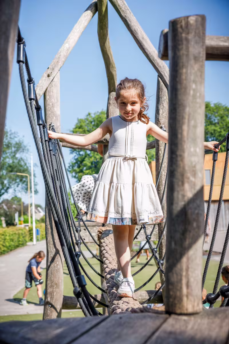 Meisje in witte jurk loopt op touwbrug in speeltuin op een zonnige dag.