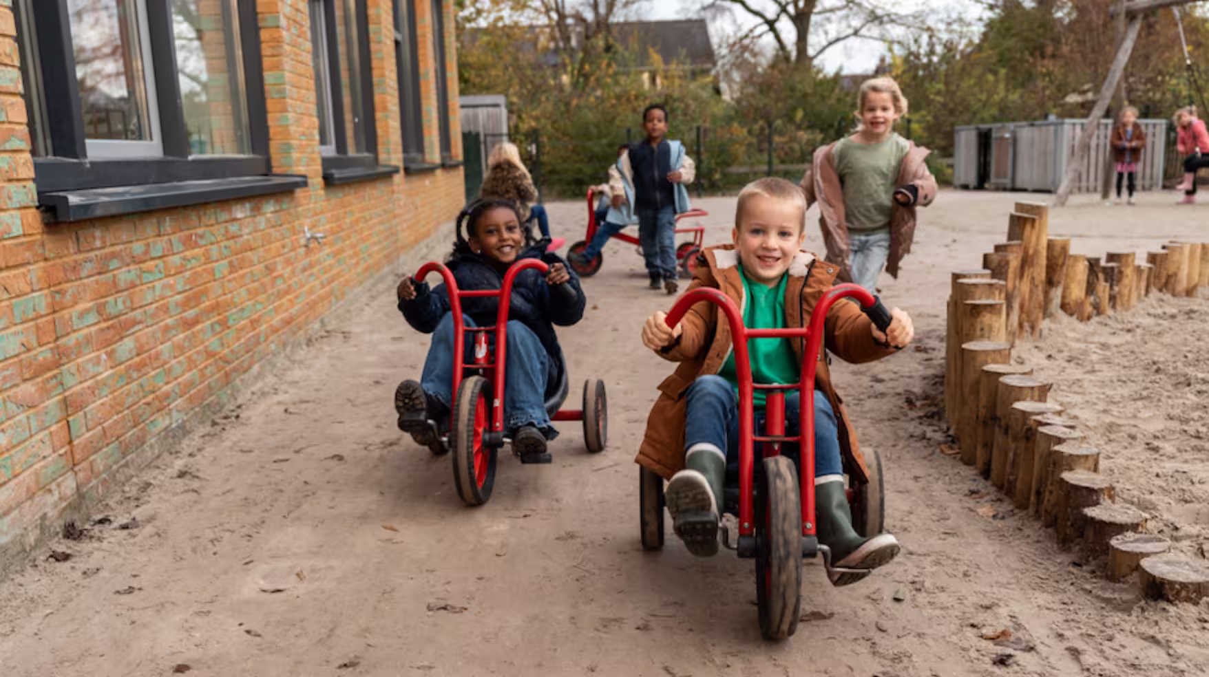 Kinderen spelen buiten met rode driewielers op een zandpad naast een bakstenen gebouw.
