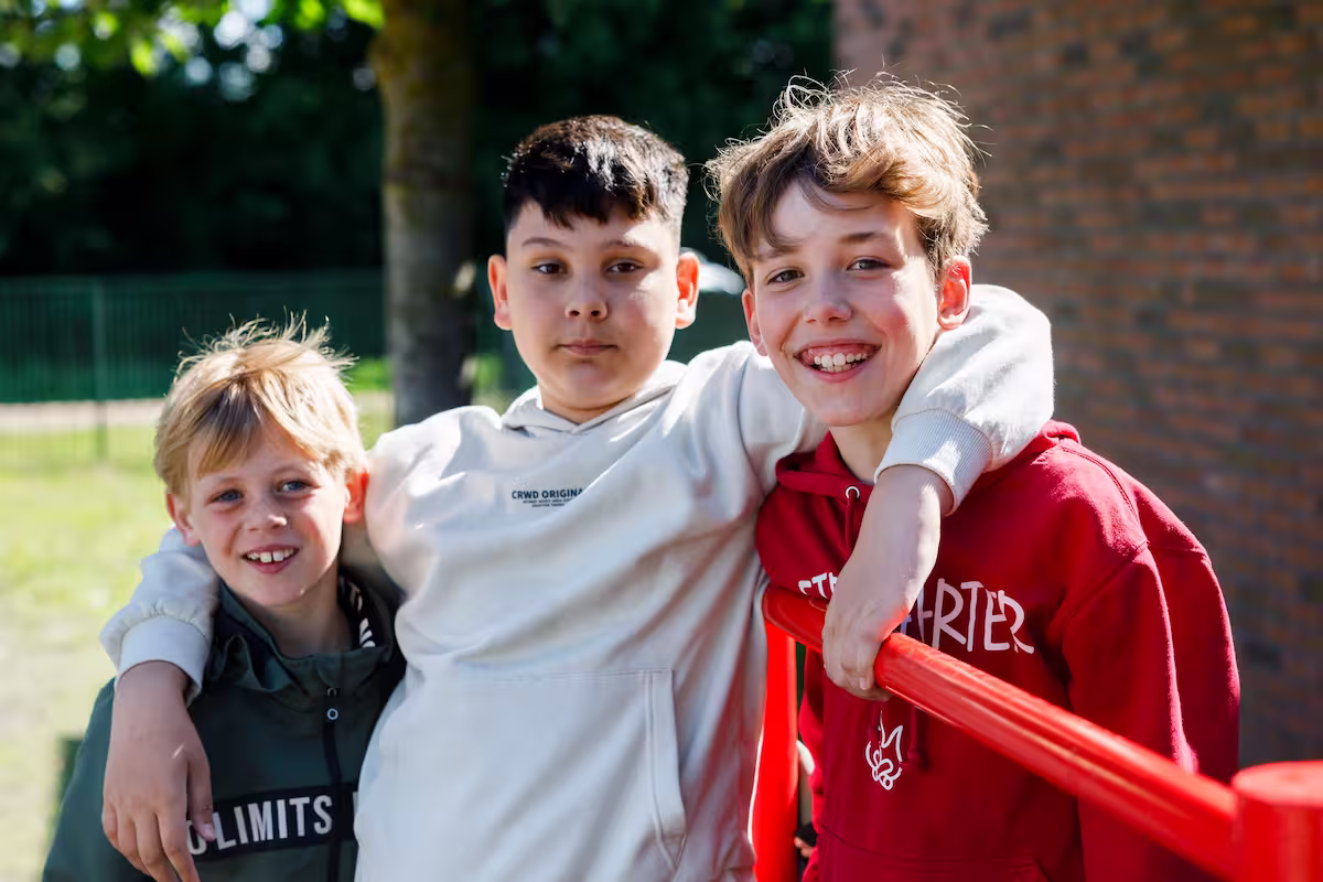 Drie jongens buiten met hun armen om elkaar, lachend bij een rood hek onder een boom.