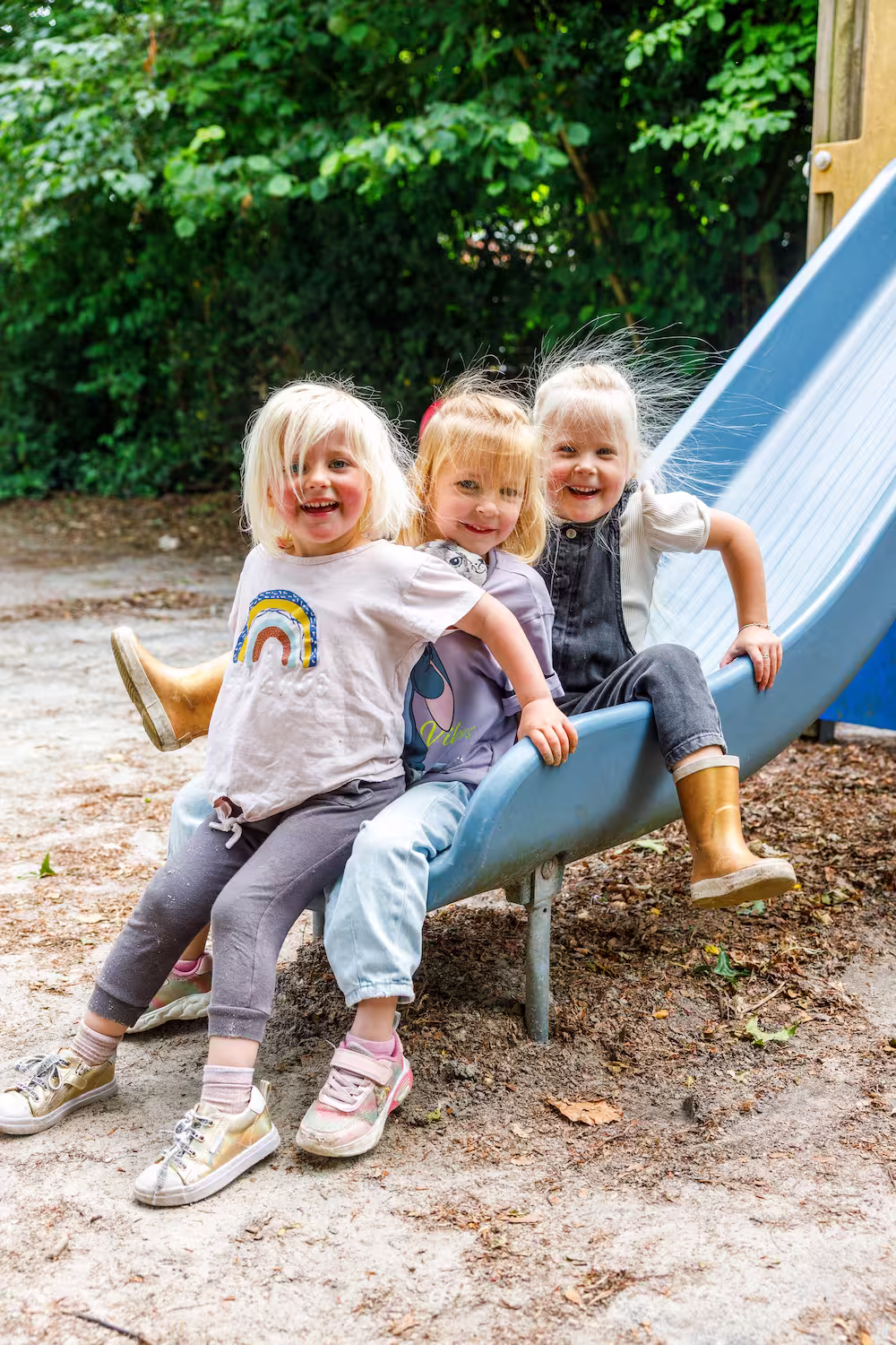 Drie lachende kinderen zitten samen aan het einde van een glijbaan in een speeltuin.