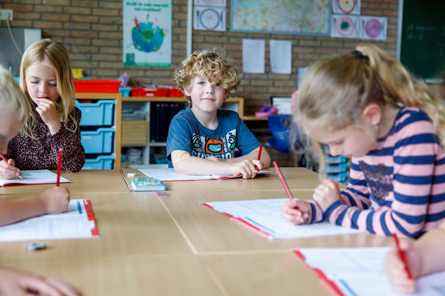 Vier jonge kinderen zitten aan een tafel in een klaslokaal en schrijven in werkboeken met rode potloden.