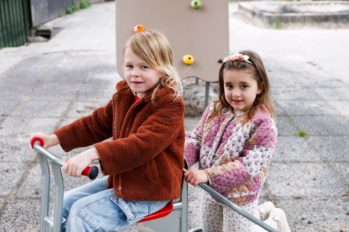 Twee kinderen spelen buiten, een zit op een speelapparaat en de ander duwt het.