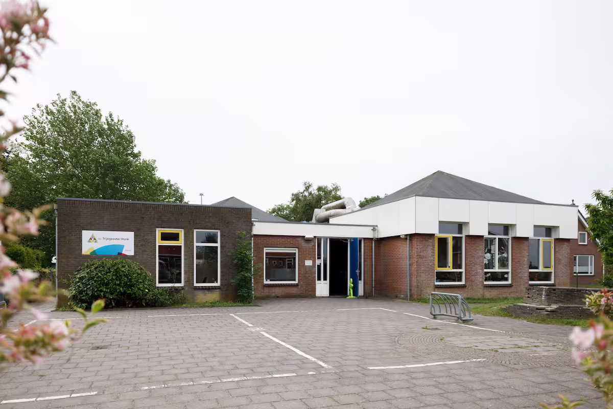Schoolgebouw met bakstenen gevel, ramen met gele kozijnen en een open ingang omgeven door bomen.