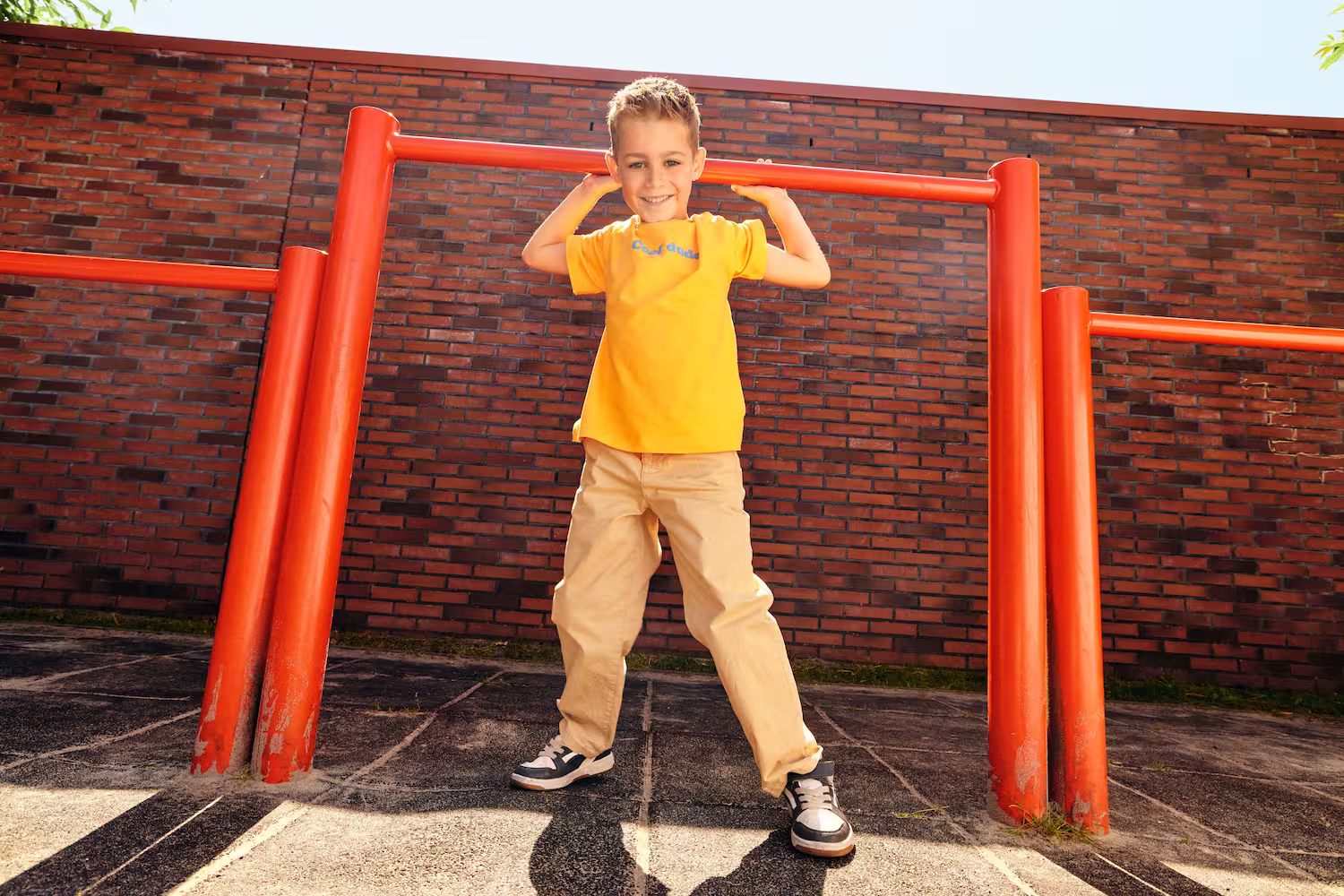 Jongen doet pull-up aan feloranje optrekstang buiten, lachend met een geel T-shirt en beige broek.