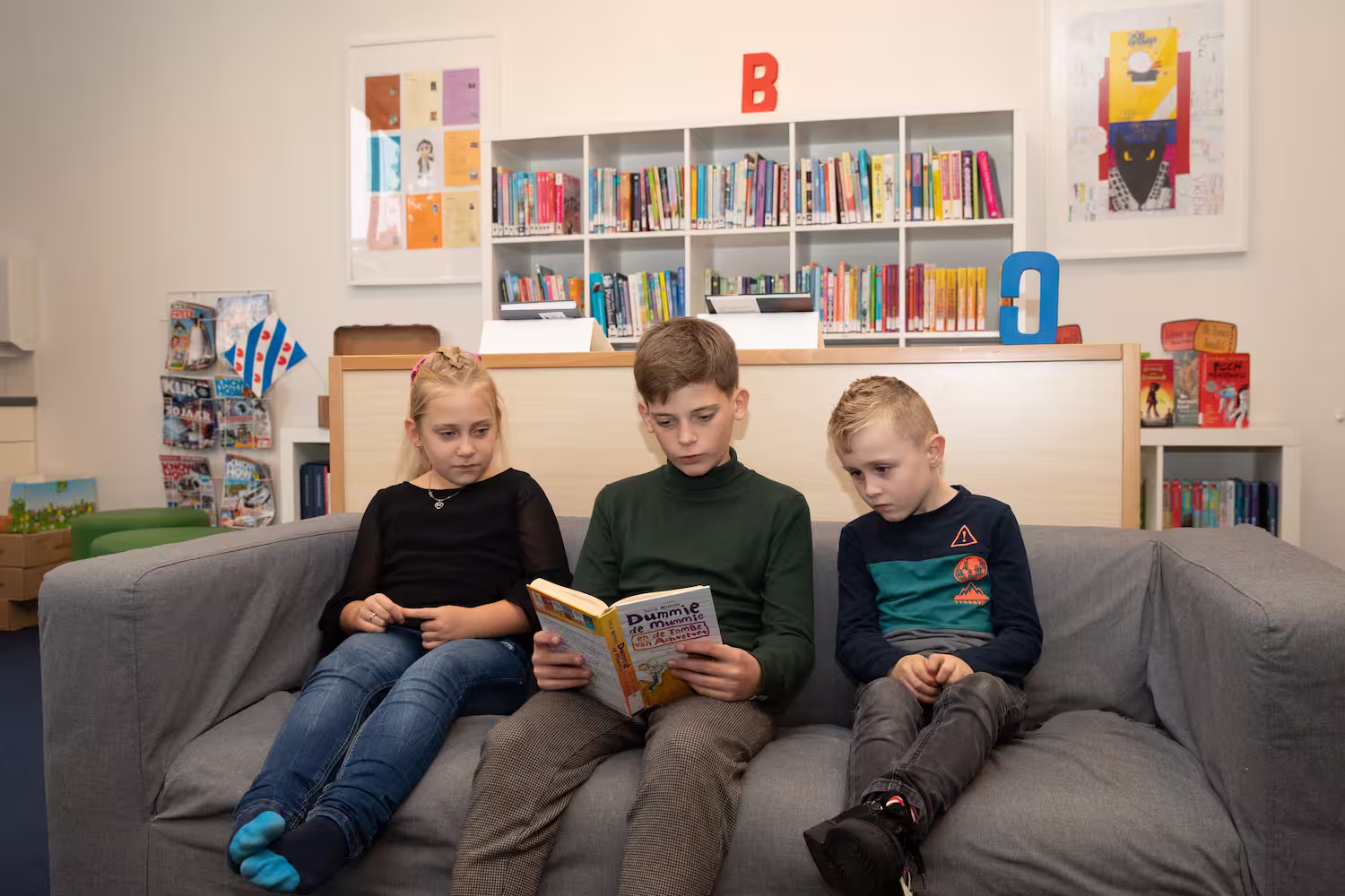 Drie kinderen zitten op een bank in een bibliotheek terwijl de middelste een boek leest.