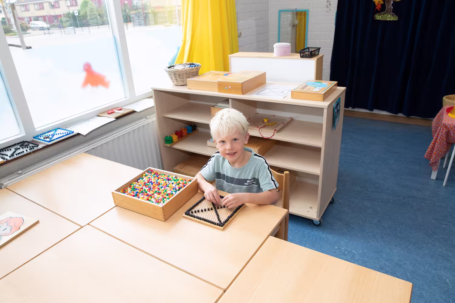 Blond jongetje zittend aan tafel met kralen en een kralenbord in een klaslokaal.