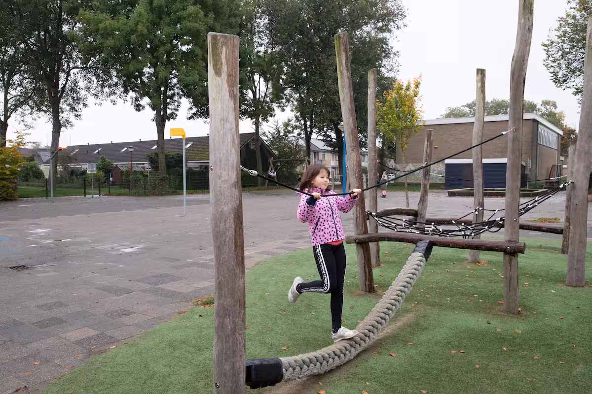 Meisje klimt op touwbrug speeltuin omringd door houten palen en bomen buiten.