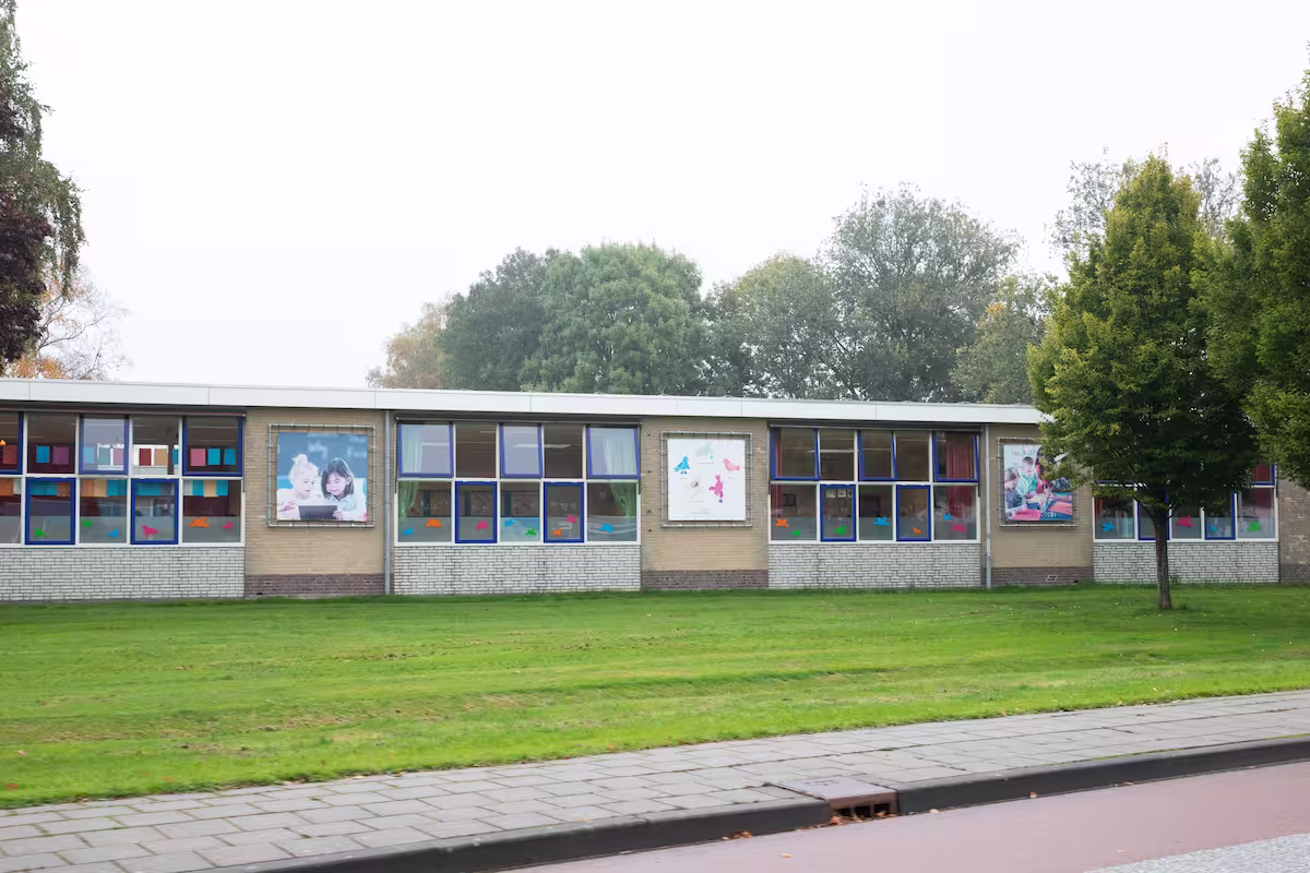 Schoolgebouw met grote ramen en kleurrijke raamdecoraties naast een fietspad en grasveld.