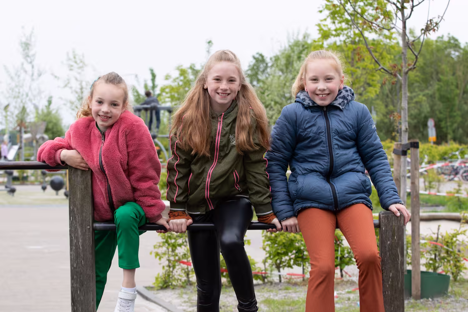 Drie lachende meisjes zitten buiten op een metalen balk in een park met bomen en planten.