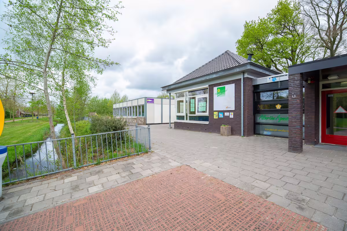 Modern schoolgebouw naast een begroeide waterloop met bomen en een betegeld plein ervoor.