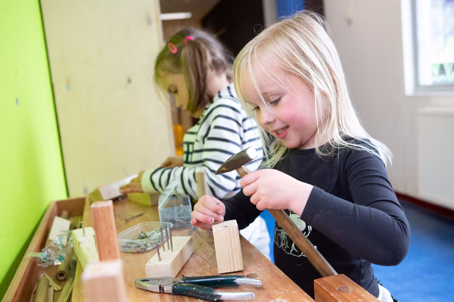 Twee meisjes werken met hout en gereedschap, een gebruikt een hamer om een spijker in hout te slaan.