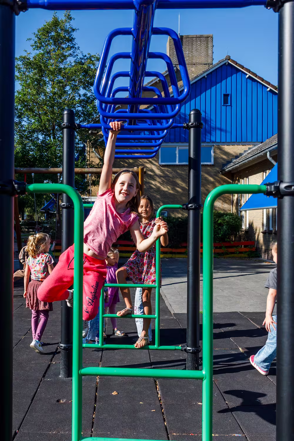 Kinderen spelen op een klimrek met blauwe stangen op een schoolplein buiten bij een gebouw.