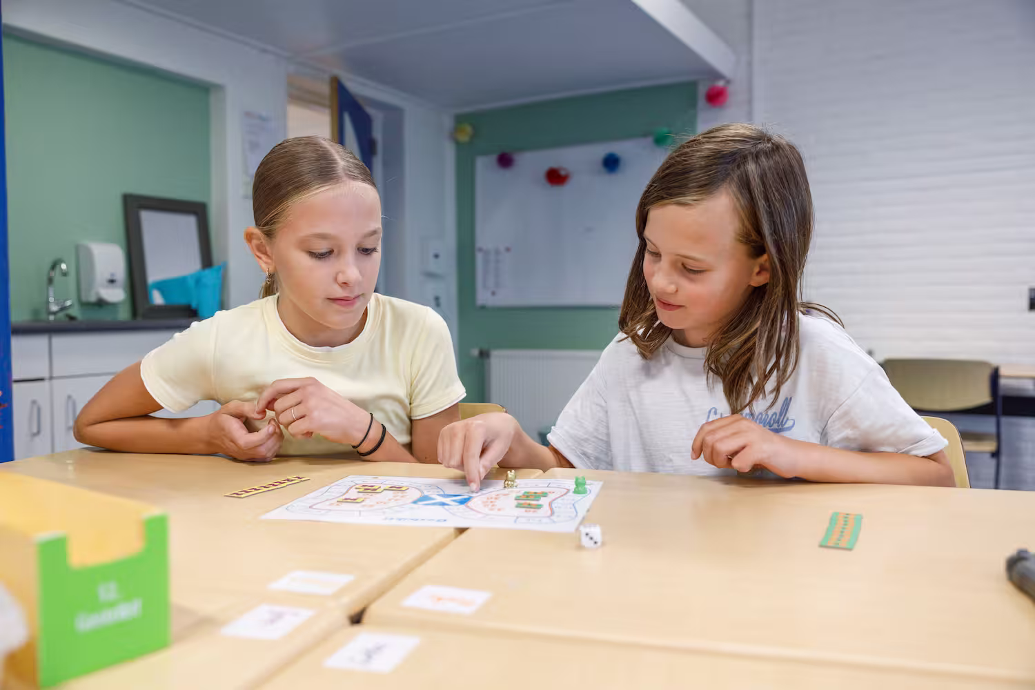 Twee meisjes spelen een bordspel aan een tafel in een klaslokaal.