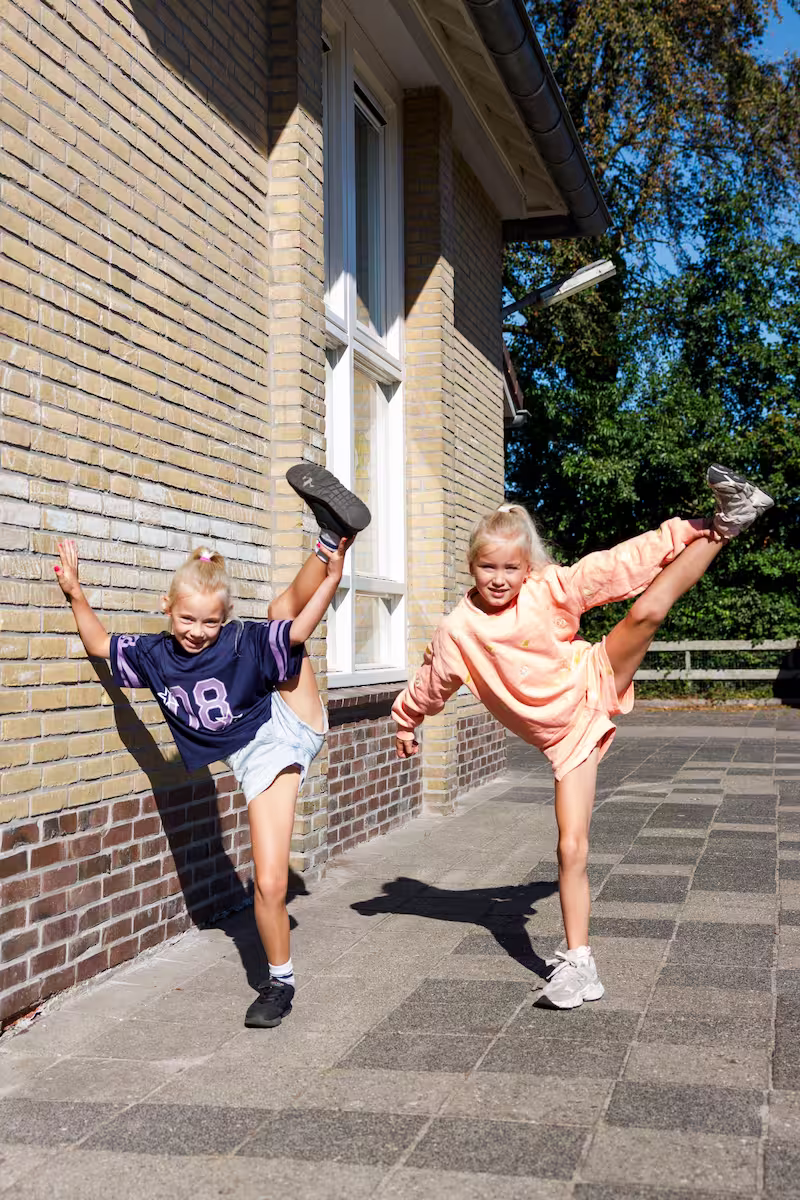 Twee blonde meisjes strekken een been omhoog terwijl ze buiten naast een bakstenen huis staan.