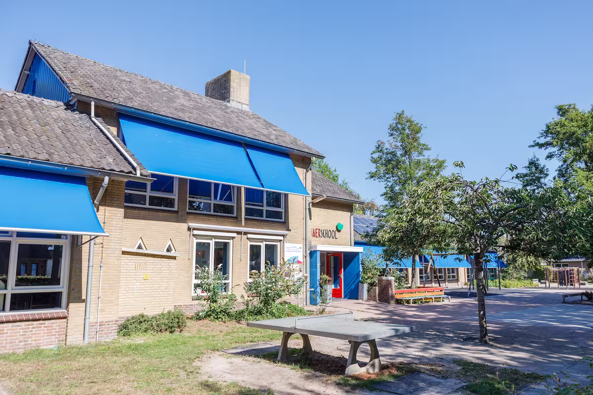 Schoolgebouw met blauwe zonneschermen, een boom en een tafeltennistafel op het schoolplein.