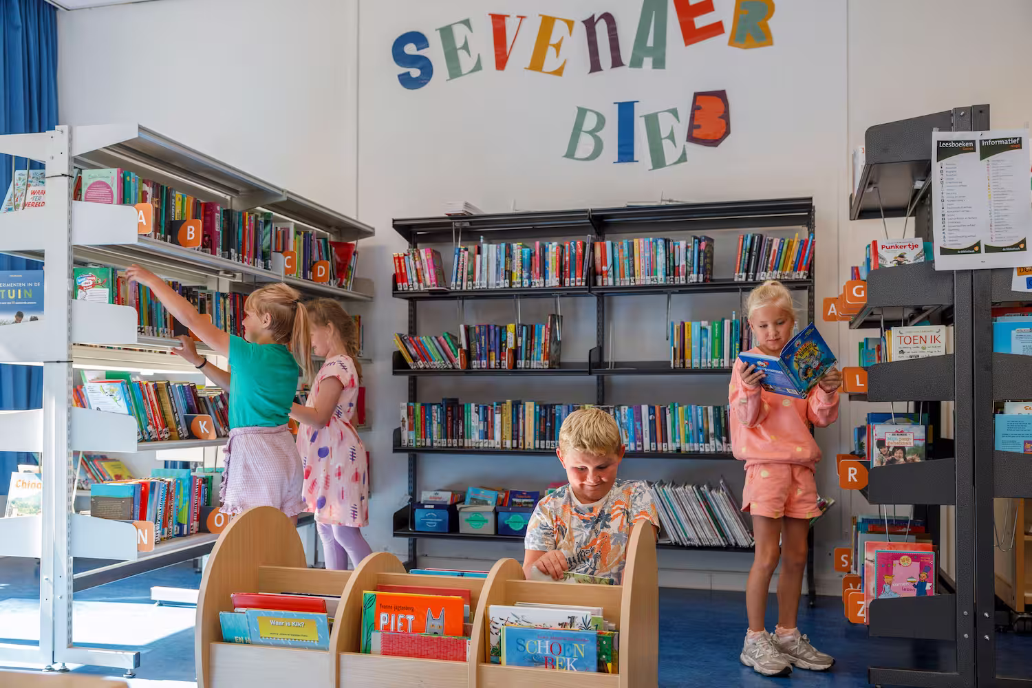 Vier kinderen lezen en kiezen boeken in een kleurrijke bibliotheek met alfabetische labels.