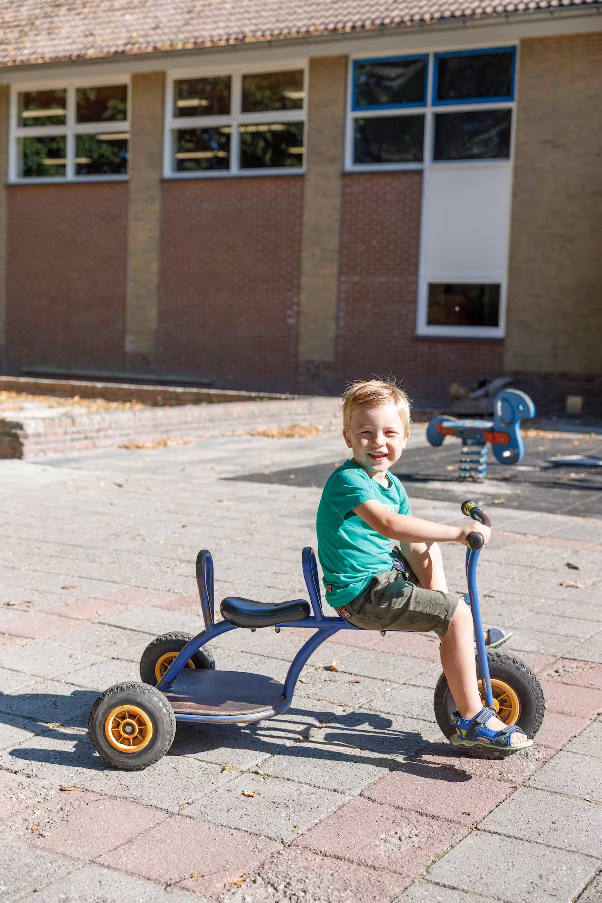 Jongen zittend op een blauw driewielig fietsje buiten in zonlicht bij een gebouw.
