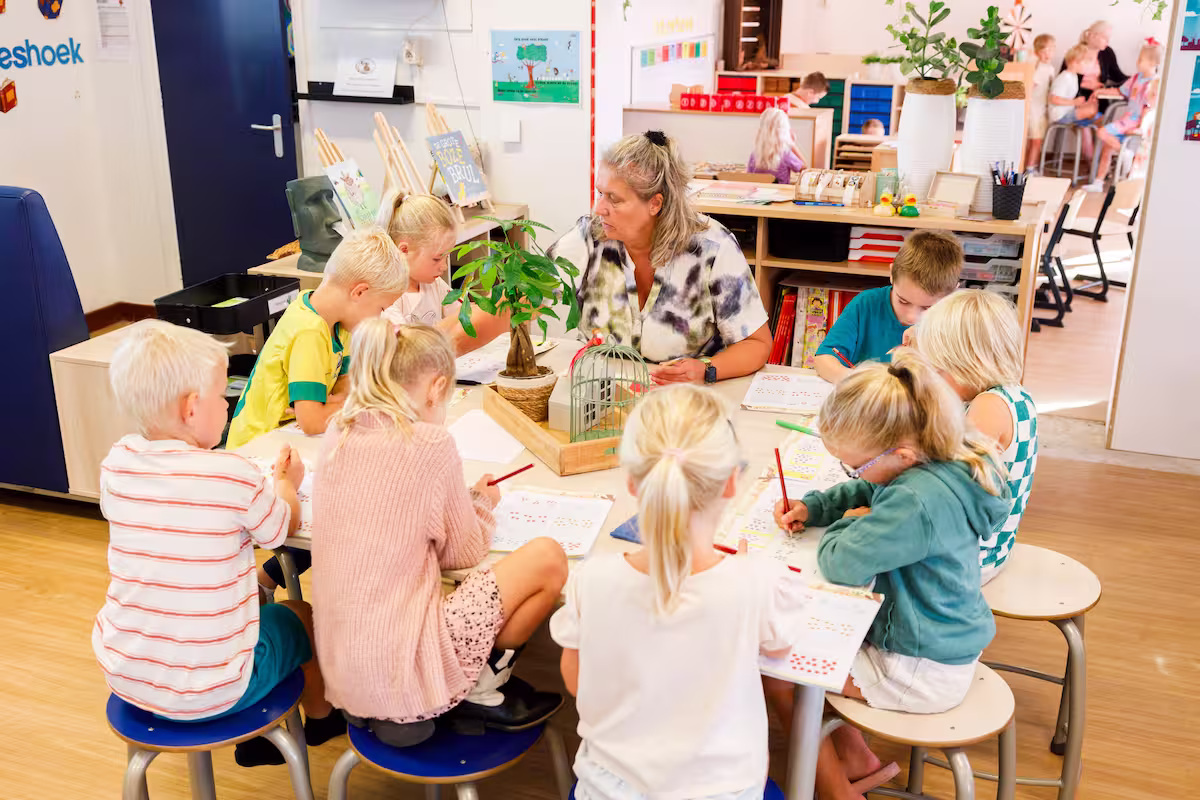 Juf helpt acht jonge kinderen met kleurplaten in een klaslokaal met tafels en stoelen.