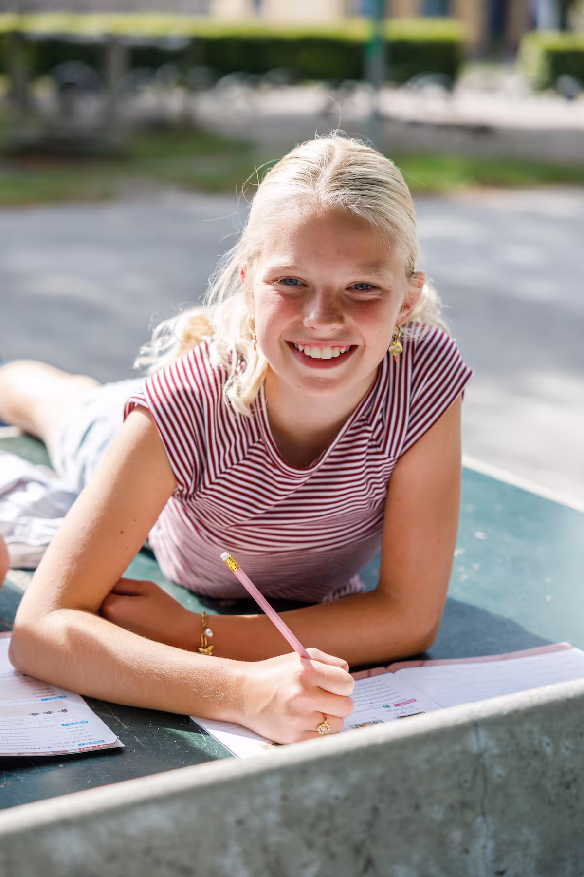 Blond meisje buiten liggend op tafel, schrijvend in notitieboek met potlood en lachend.