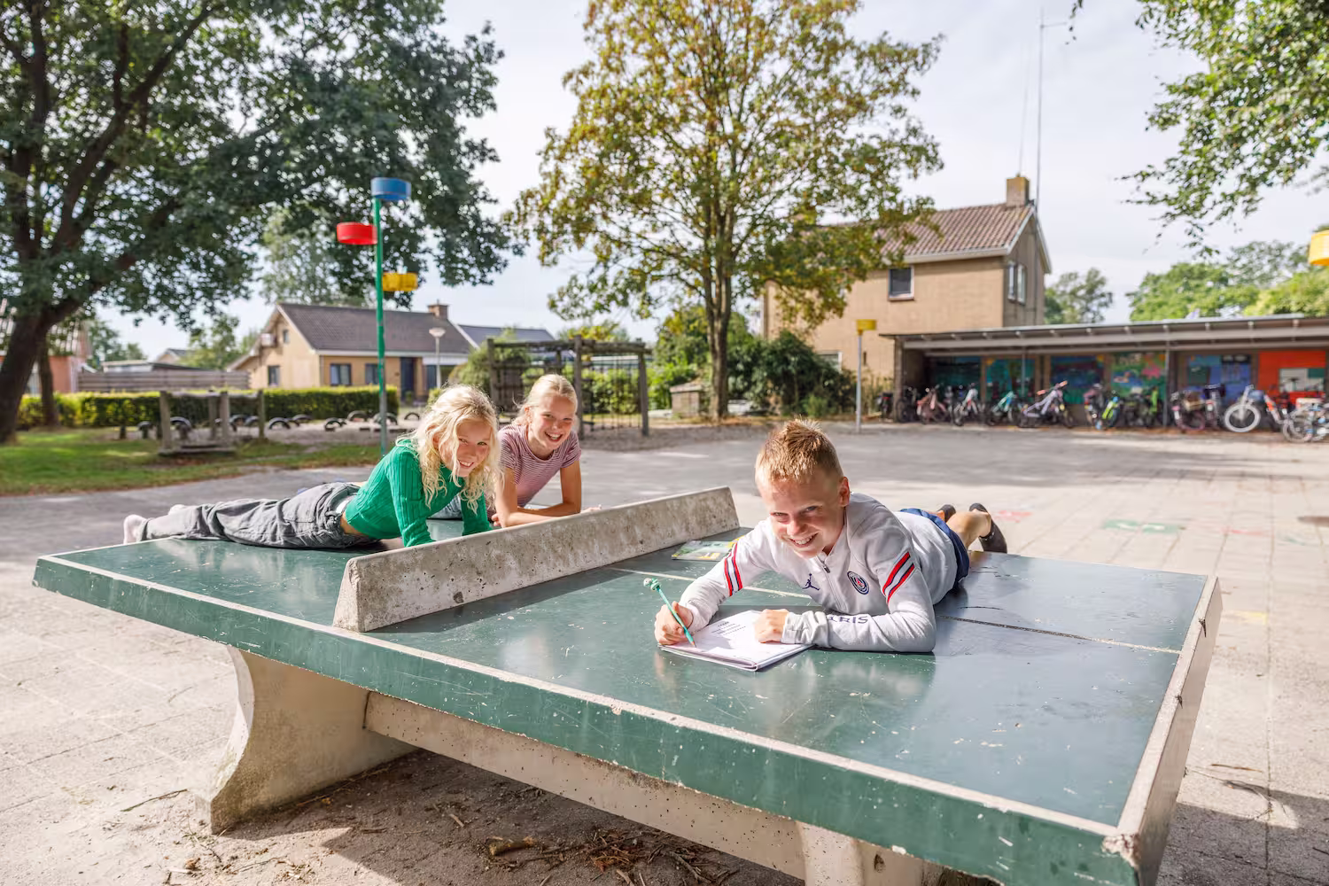 Drie lachende kinderen liggen op een tafeltennistafel buiten, een jongen schrijft in een schrift.
