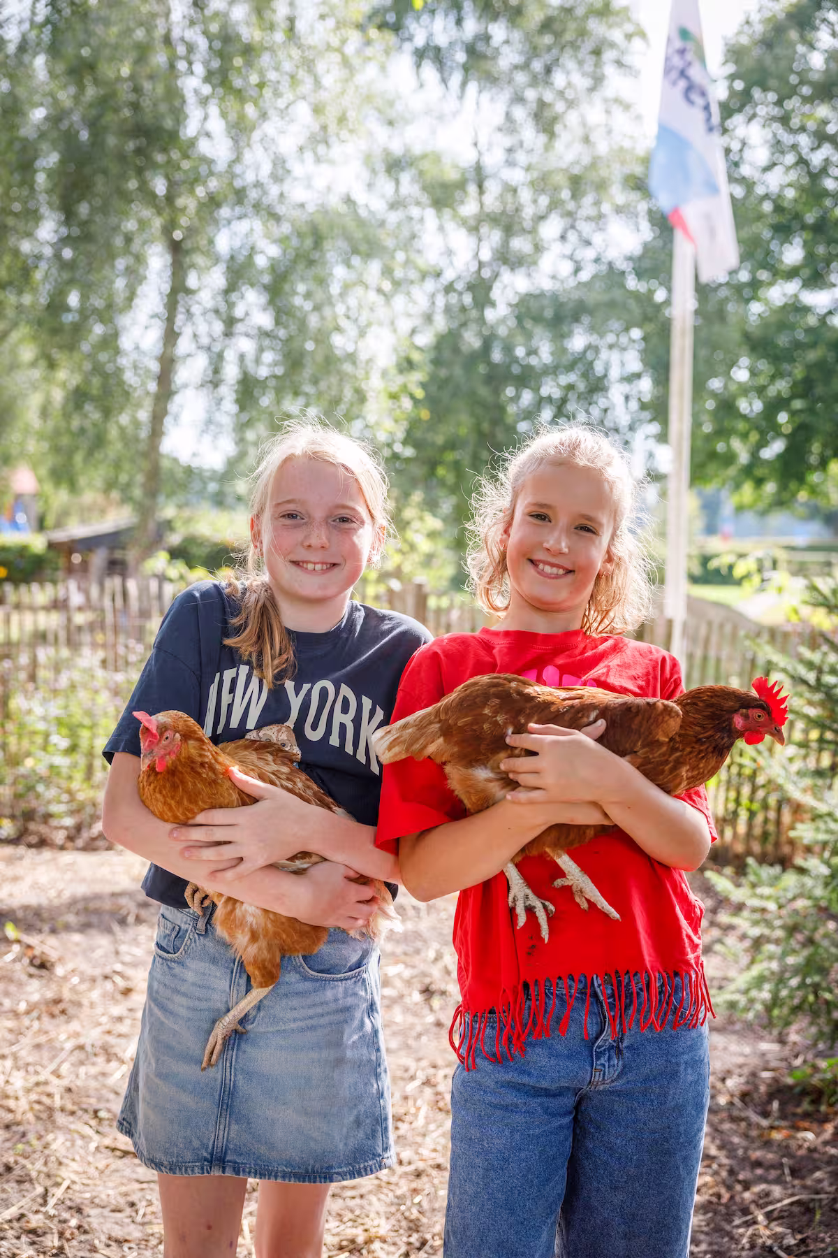 Twee meisjes lachen en houden elk een bruine kip vast in een zonnige buitenomgeving.