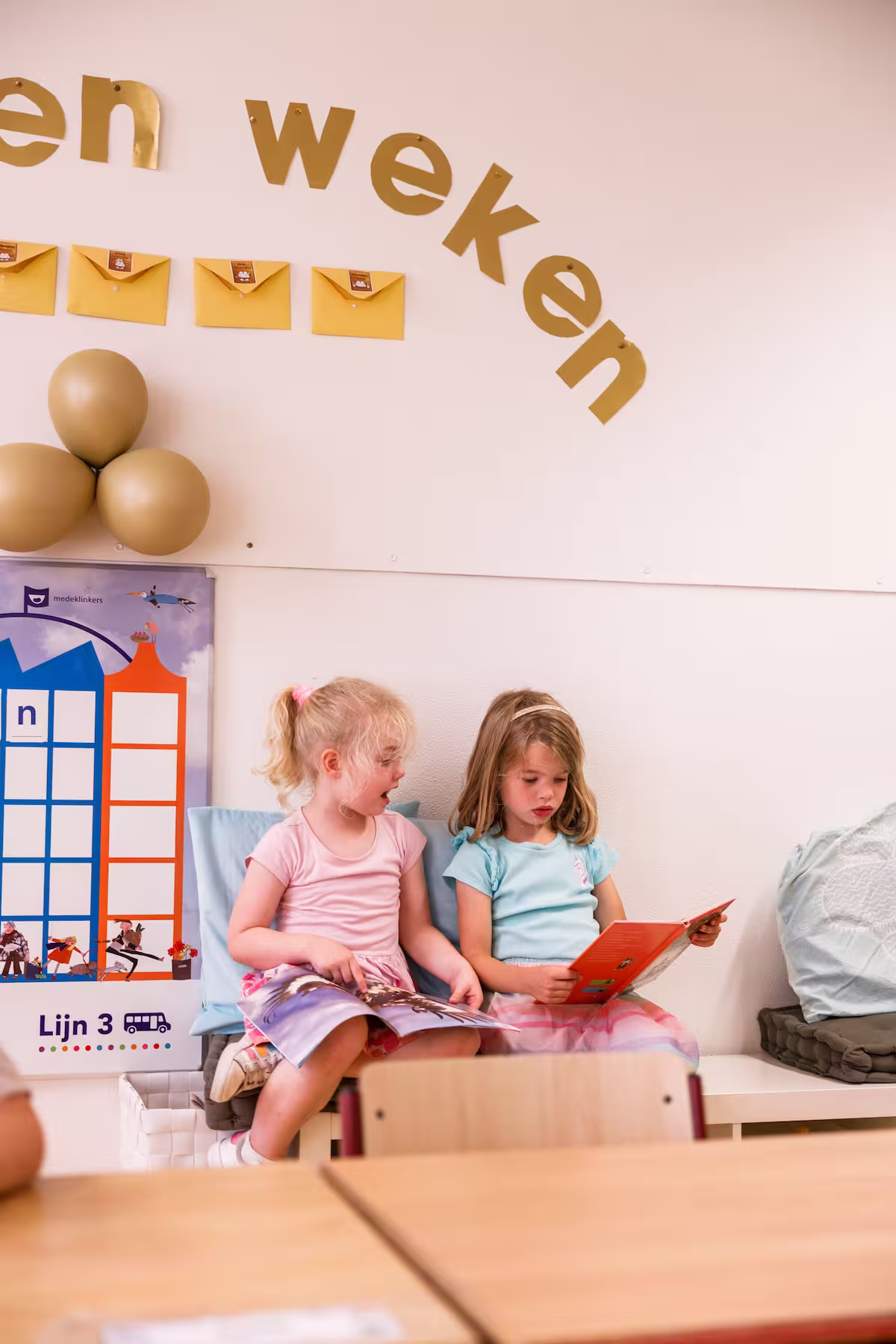 Twee jonge meisjes zitten op een bankje en lezen boeken in een klaslokaal met wanddecoraties.