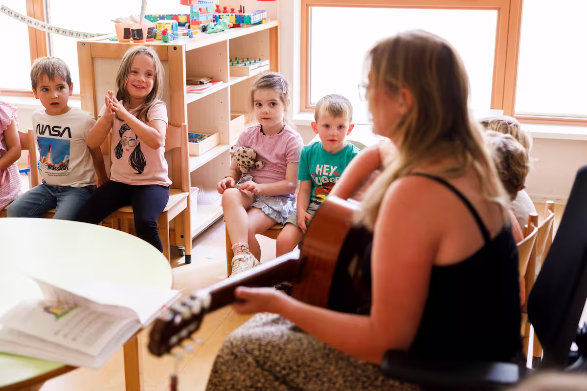 Vrouw speelt gitaar voor zittende kinderen in klaslokaal met speelgoed en ramen.