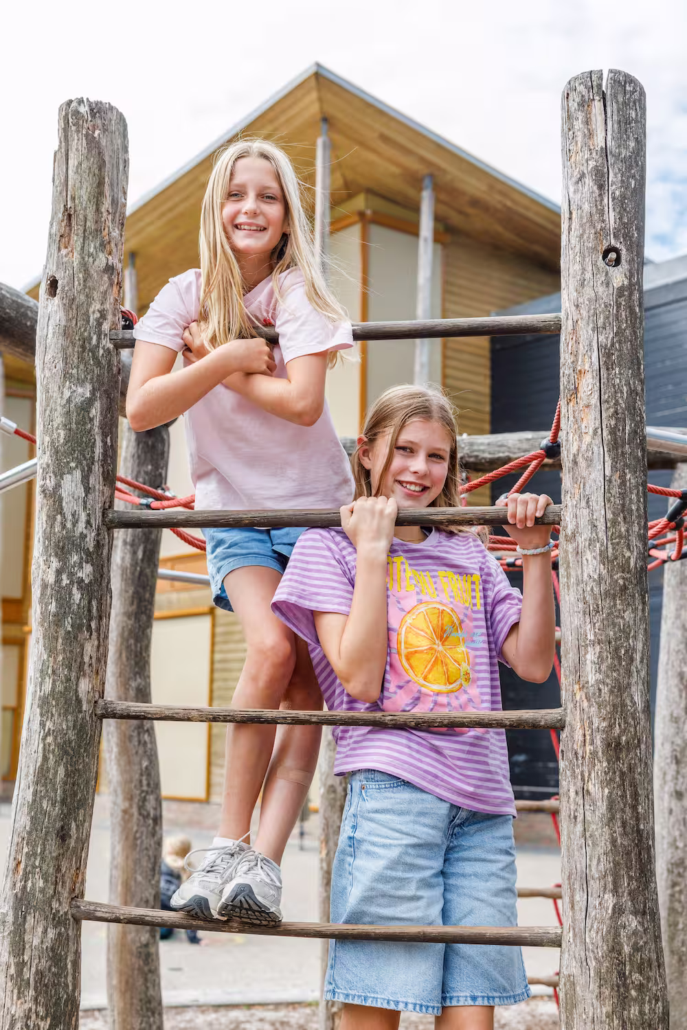 Twee lachende meisjes klimmen op een houten klimrek buiten bij een moderne school.