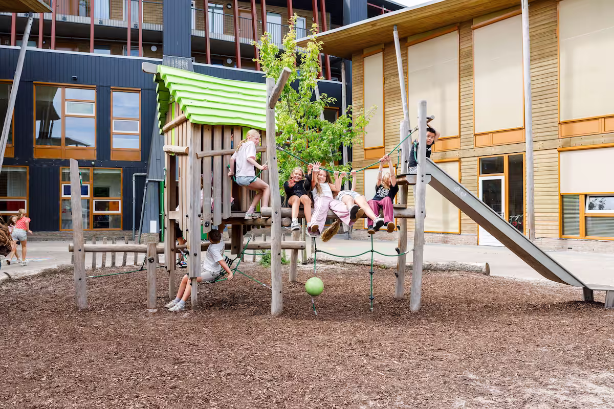 Kinderen spelen op houten speeltoestellen met glijbaan en klimnet bij schoolgebouw.