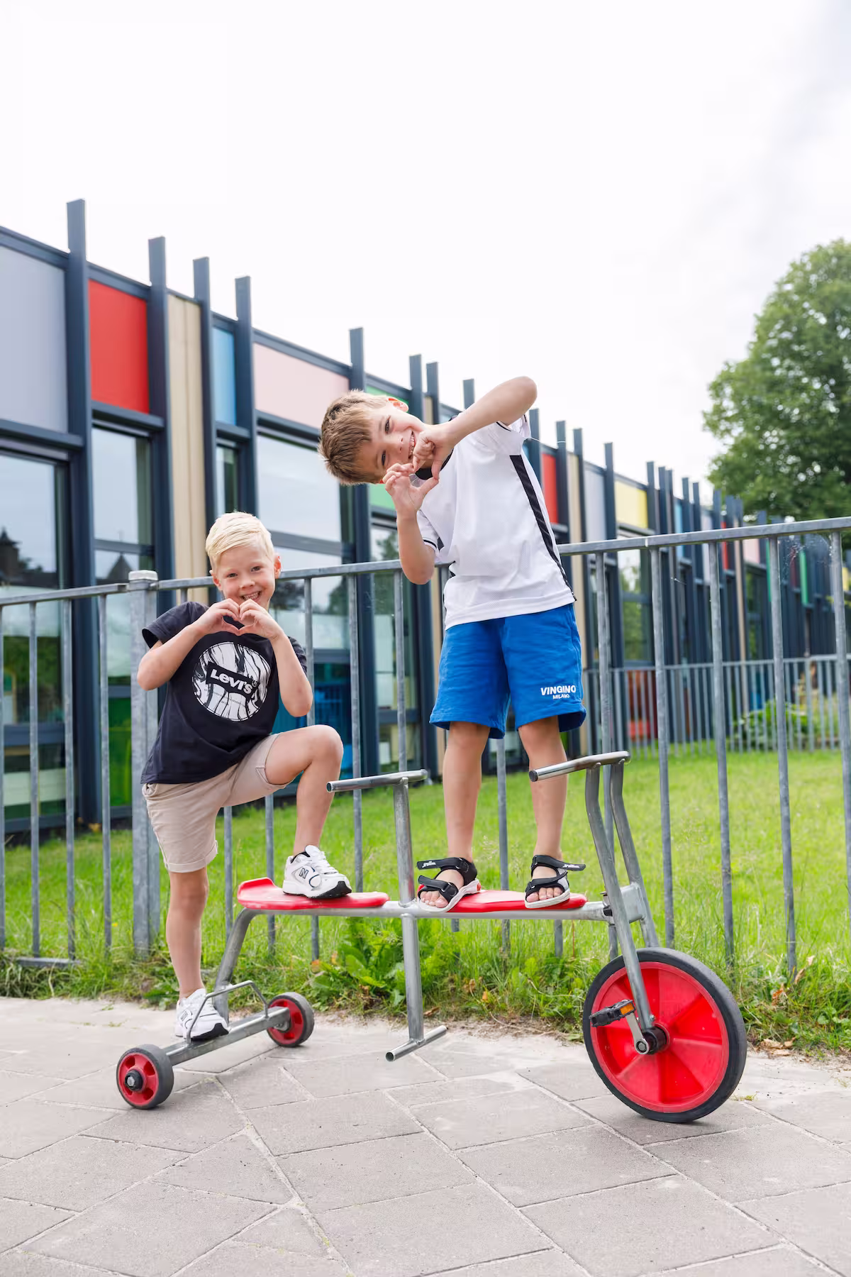Twee jongens staan buiten met een driewielige duofiets en maken hartvormen met hun handen.