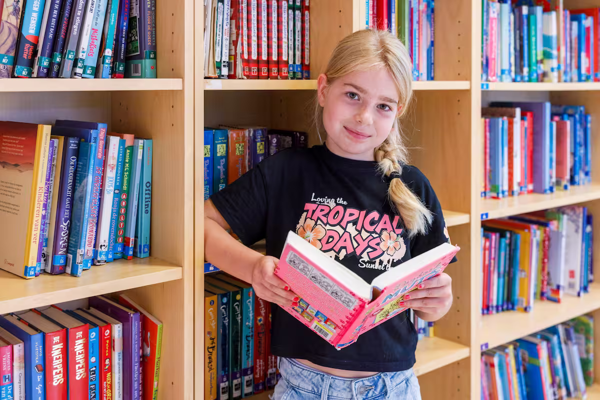 Jong meisje met blonde vlecht leest een boek in een bibliotheek met houten boekenplanken.